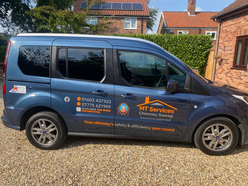 A blue van with "MT Services Chimney Sweep" branding, contact details, and business logos parked on a gravel drive in front of a brick house.