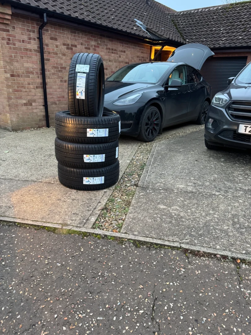 Four new car tyres are stacked on a driveway in front of a house, with a black electric car nearby and its front boot open.