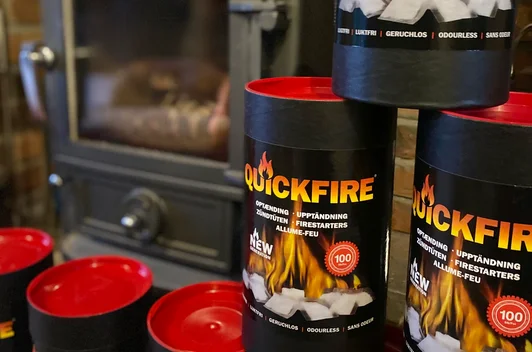 Three containers of Quickfire firelighters are displayed in front of a wood-burning stove, with their lids removed and stacked nearby.